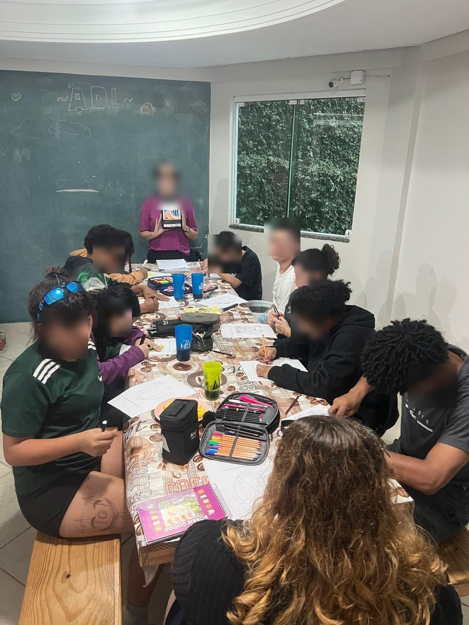 Adolescentes participam de oficina de desenho em volta de uma mesa com lápis de cor, com rostos desfocados para preservar a identidade