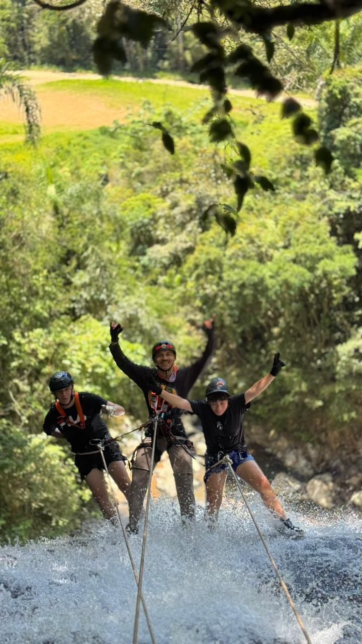 Participantes com capacete e equipamentos de segurança descem a cachoeira em rapel acompanhados por instrutores