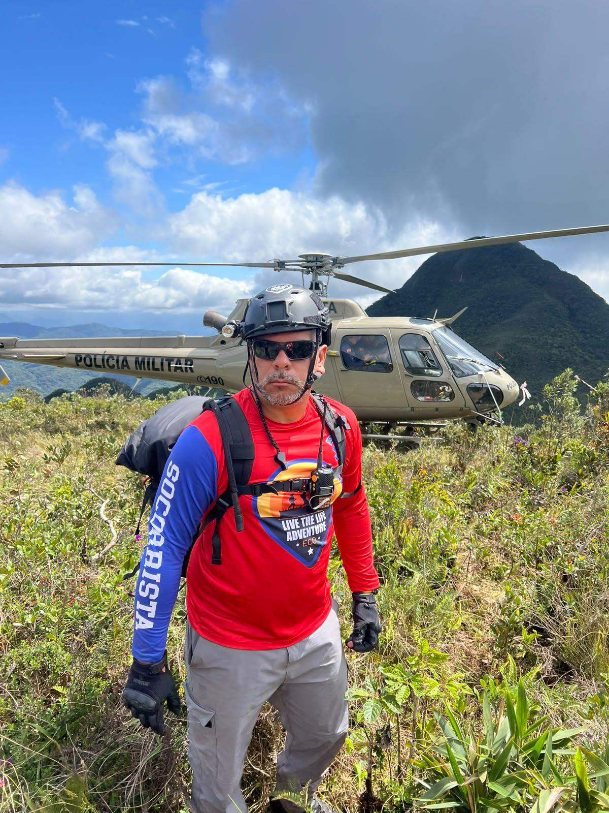 Socorrista da associação em operação de busca em montanha, com helicóptero da Polícia Militar ao fundo